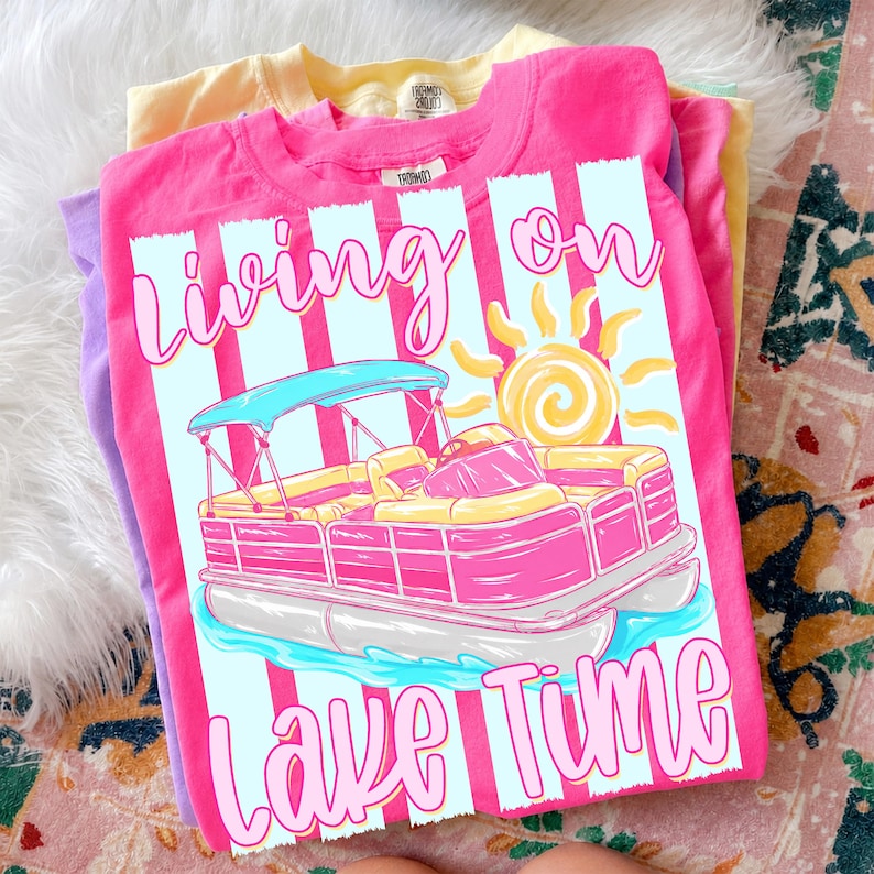 May include: A stack of three t-shirts in pink, yellow, and purple. The pink shirt features a graphic of a pink and yellow pontoon boat with the text "Living on Lake Time". The design includes a sun and blue stripes.