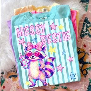May include: A stack of colorful t-shirts. The top shirt is light blue with the words "MESSY BESTIE" and a cartoon raccoon wearing heart-shaped sunglasses and holding a drink. Pink, yellow, and purple shirts are visible underneath.