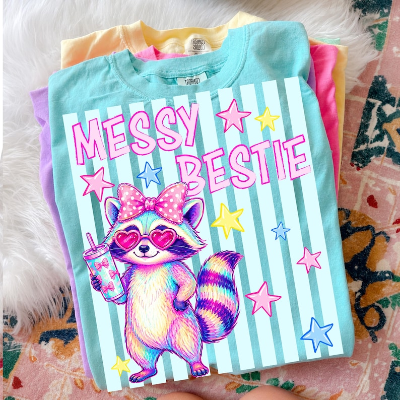 May include: A stack of colorful t-shirts. The top shirt is light blue with the words "MESSY BESTIE" and a cartoon raccoon wearing heart-shaped sunglasses and holding a drink. Pink, yellow, and purple shirts are visible underneath.