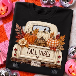May include: A black t-shirt featuring a graphic of a cream-colored pickup truck carrying various pumpkins, autumn leaves, and a flower. The truck tailgate displays the text FALL VIBES with a small heart and a license plate reading PUMPKIN.