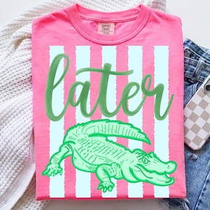 Peut inclure: T-shirt rose avec un motif d'alligator vert et le mot "later" en vert. Le t-shirt présente des rayures verticales bleu clair et une étiquette Comfort Colors. Le t-shirt est associé à un jean bleu et à une coque de téléphone.