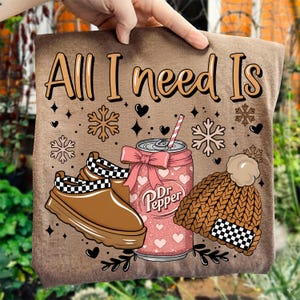 May include: A brown t-shirt with the text "All I need Is" in a decorative font. The design includes a brown shoe with a black and white checkered pattern, a Dr. Pepper can with a pink bow, and a brown knit hat with a checkered pattern.