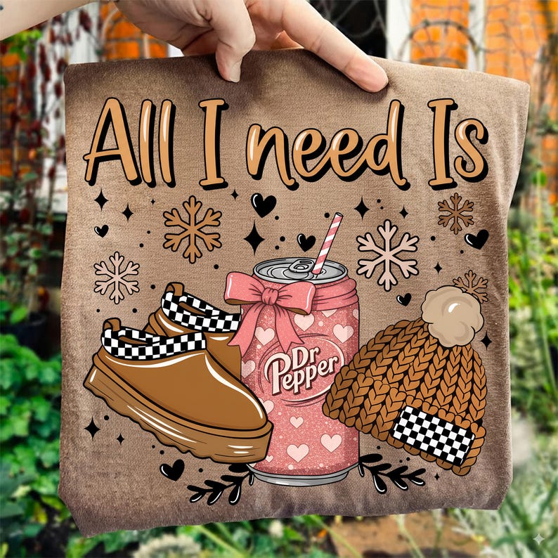 May include: A brown t-shirt with the text "All I need Is" in a decorative font. The design includes a brown shoe with a black and white checkered pattern, a Dr. Pepper can with a pink bow, and a brown knit hat with a checkered pattern.
