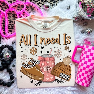 May include: Cream-colored t-shirt with the text "All I need Is" above a graphic of a Dr. Pepper can, shoes, and a beanie. The can is pink with hearts, the shoes are brown and black, and the beanie is brown with a checkered pattern.