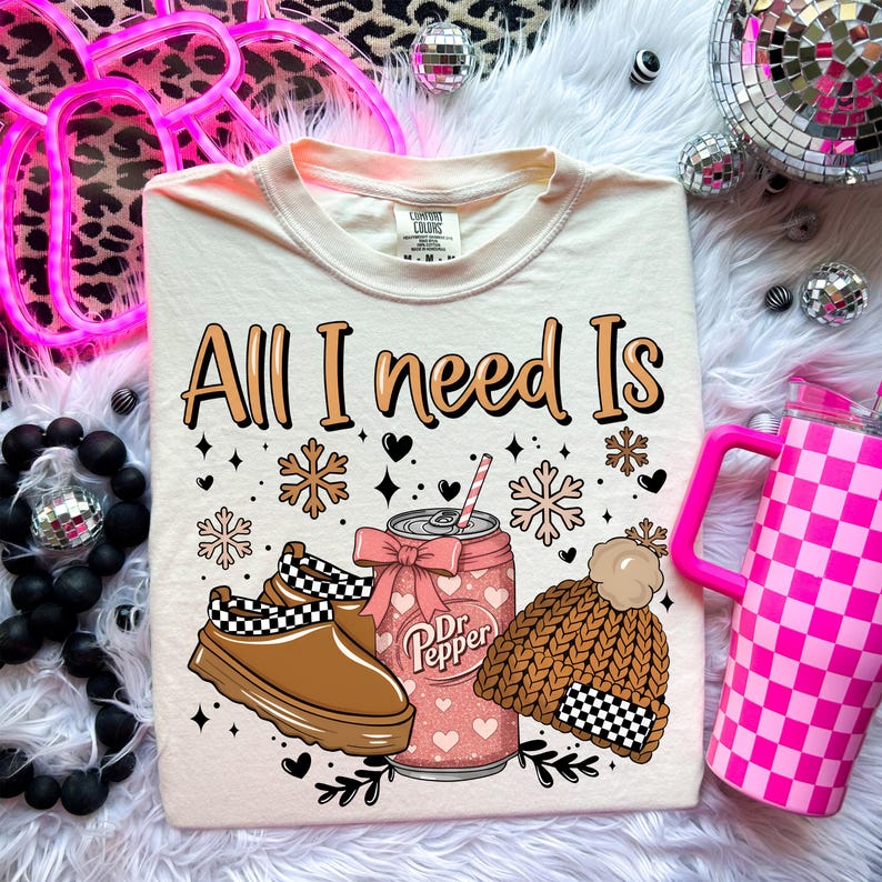 May include: Cream-colored t-shirt with the text "All I need Is" above a graphic of a Dr. Pepper can, shoes, and a beanie. The can is pink with hearts, the shoes are brown and black, and the beanie is brown with a checkered pattern.