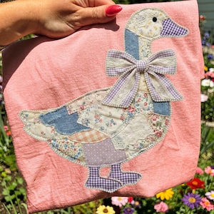 May include: A pink t-shirt featuring a patchwork goose design. The goose has a blue denim neck, a gingham beak, and a bow with lace trim. The body is made of floral and patterned fabrics, with a dotted purple patch.