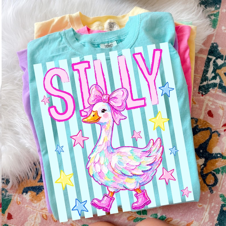 May include: A stack of pastel-colored t-shirts. The top shirt is light blue with the word "SILLY" in pink, a cartoon goose wearing a bow and boots, and star accents. Other shirts are yellow, pink, and purple.