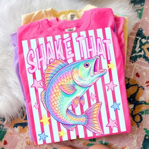 May include: A stack of colorful t-shirts, including pink, yellow, and purple. The top shirt is pink and features a graphic of a rainbow-colored fish with the text "SHAKE THAT" in white. The design includes stars and vertical stripes.