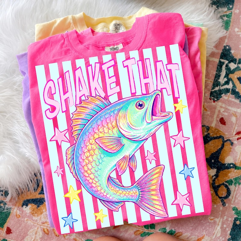 May include: A stack of colorful t-shirts, including pink, yellow, and purple. The top shirt is pink and features a graphic of a rainbow-colored fish with the text "SHAKE THAT" in white. The design includes stars and vertical stripes.