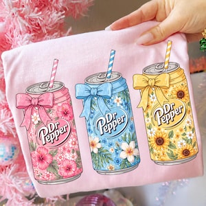 以下が含まれることがあります： ピンク色のTシャツに、3つの様式化されたDr. Pepperのソーダ缶が描かれています。各缶は、花の模様、リボン、ストローで装飾されています。缶はピンク、青、黄色で、Dr. Pepperのロゴが大きく表示されています。