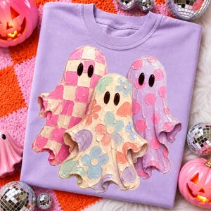 Pu&ograve; includere: Una t-shirt color viola chiaro con una grafica raffigurante tre fantasmi. Il fantasma a sinistra ha un motivo a scacchi rosa e bianco, quello centrale un design floreale multicolore e quello a destra un motivo a pois viola e rosa.