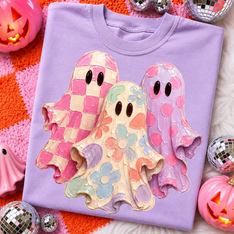 Pu&ograve; includere: Una t-shirt color viola chiaro con una grafica raffigurante tre fantasmi. Il fantasma a sinistra ha un motivo a scacchi rosa e bianco, quello centrale un design floreale multicolore e quello a destra un motivo a pois viola e rosa.