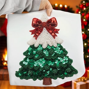 May include: White cushion with a sequined Christmas tree design. The tree features a red sequined bow, white beaded snow, and green sequined branches. The tree trunk is brown.