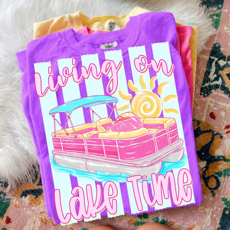 May include: A stack of colorful t-shirts, with a purple shirt on top featuring a graphic of a pink and yellow pontoon boat with the text "Living on Lake Time". The other shirts are yellow and pink.
