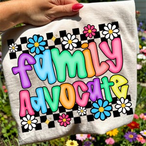 Puede incluir: Un bolso tote beige con las palabras "FAMILY ADVOCATE" en letras de burbujas coloridas. El diseño incluye un patrón de cuadros blancos y negros, margaritas y detalles de estrellas. El bolso es sostenido por una mano.
