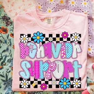 May include: A light pink t-shirt with the words "Behavior Support" in a colorful, patterned design. The text is outlined in black and surrounded by flowers and a checkered pattern. The shirt is made of a soft material.