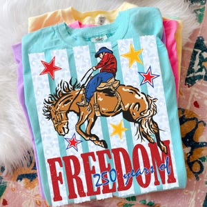 May include: A light blue t-shirt featuring a graphic of a cowboy riding a bucking horse. The design includes red, white, and blue stars, vertical stripes, and the red text "FREEDOM" with the blue text "250 years of" layered over the bottom.
