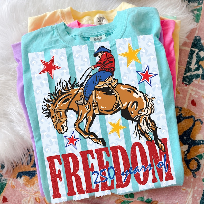 May include: A light blue t-shirt featuring a graphic of a cowboy riding a bucking horse. The design includes red, white, and blue stars, vertical stripes, and the red text "FREEDOM" with the blue text "250 years of" layered over the bottom.