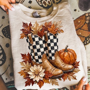 Puede incluir: Una sudadera beige de cuello redondo con un estampado gráfico de dos botas vaqueras marrones con un patrón de cuadros blancos y negros. Las botas están rodeadas de hojas de otoño naranjas y marrones, una flor blanca y una calabaza naranja.