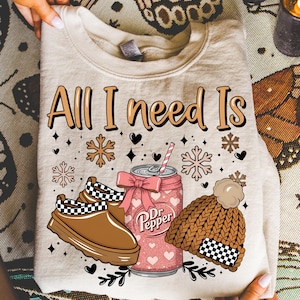 May include: A beige sweatshirt with the text "All I need Is" and winter-themed graphics. The design includes a Dr. Pepper can with a bow, a pair of brown clogs, and a brown knit hat with a pom-pom.
