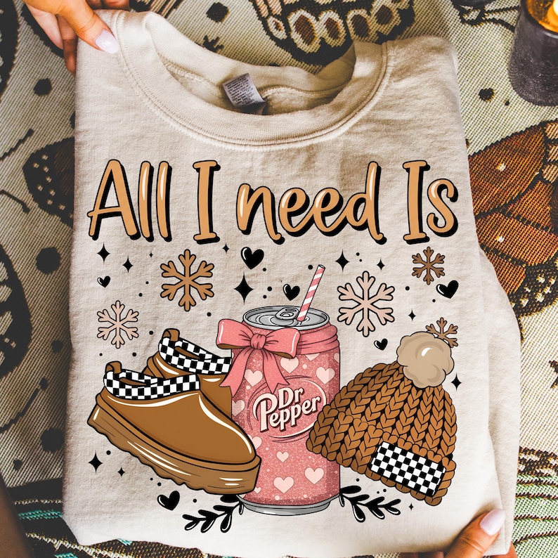 May include: A beige sweatshirt with the text "All I need Is" and winter-themed graphics. The design includes a Dr. Pepper can with a bow, a pair of brown clogs, and a brown knit hat with a pom-pom.