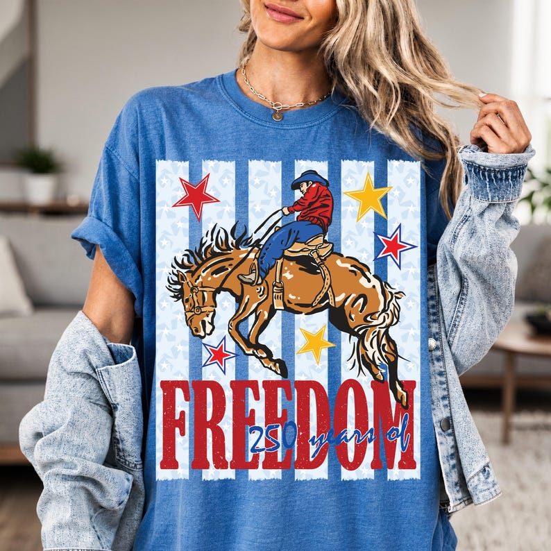 May include: A blue t-shirt featuring a graphic of a cowboy riding a bucking horse. The design includes vertical stripes, red and yellow stars, and the red text "FREEDOM" with "250 years of" written in blue script below the main word.