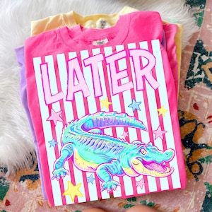 May include: A stack of three t-shirts in yellow, purple, and pink. The top pink shirt features a graphic of an alligator with the word "LATER" in pink letters. The alligator is in shades of blue, green, and yellow, with star accents.