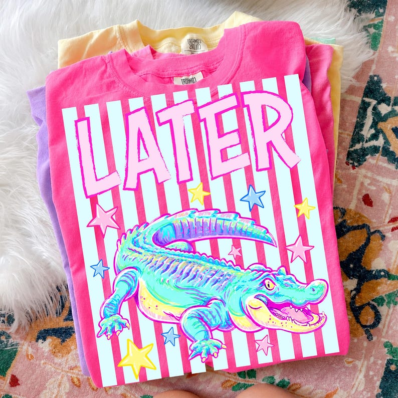 May include: A stack of three t-shirts in yellow, purple, and pink. The top pink shirt features a graphic of an alligator with the word "LATER" in pink letters. The alligator is in shades of blue, green, and yellow, with star accents.