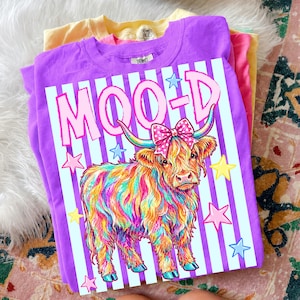 May include: A stack of colorful t-shirts. The top shirt is purple with a graphic of a Highland cow with rainbow fur, a pink bow, and the word "MOOD" in pink. The design is set against a striped background with stars.