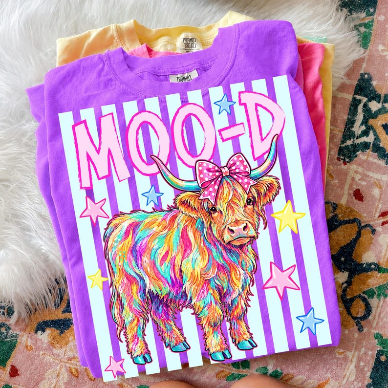May include: A stack of colorful t-shirts. The top shirt is purple with a graphic of a Highland cow with rainbow fur, a pink bow, and the word "MOOD" in pink. The design is set against a striped background with stars.