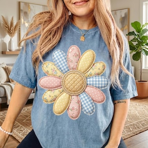 以下が含まれることがあります： 大きなカラフルなデイジーのデザインが施された、ライトブルーのTシャツ。デイジーの花びらは、ピンクのドット、黄色のドット、花柄、青のギンガムチェックなど、さまざまなパターンで構成されています。花の真ん中はキルティング加工が施されています。