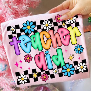 May include: A light pink t-shirt with the words "Teacher Aid" in colorful bubble letters. The shirt features a checkerboard pattern, flowers, and stars. The shirt is being held by a hand.
