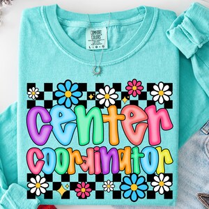 May include: A turquoise Comfort Colors t-shirt with the words "Center Coordinator" in colorful bubble letters. The design includes a black and white checkered pattern with daisy and star accents.