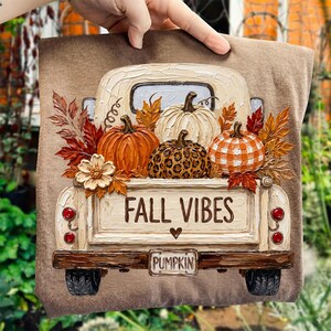 May include: A brown t-shirt featuring a graphic of a vintage truck bed filled with pumpkins, autumn leaves, and a flower. The truck tailgate displays the text FALL VIBES with a small heart, and the license plate reads PUMPKIN.