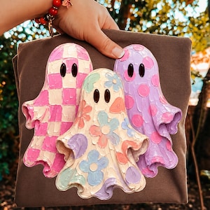 Pu&ograve; includere: Una t-shirt marrone con una grafica di tre fantasmi con effetti di pittura materica. Il fantasma a sinistra ha un motivo a scacchi rosa e bianco, quello centrale motivi floreali pastello e quello a destra pois rosa.
