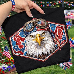 Op de afbeelding: Zwart kussen met een afbeelding van een Amerikaanse zeearend met een pilotenbril en een bandana met het ontwerp van de Amerikaanse vlag. De adelaar is geplaatst in een rood, wit en blauw zeshoekig frame. Een bijpassende armband is zichtbaar.