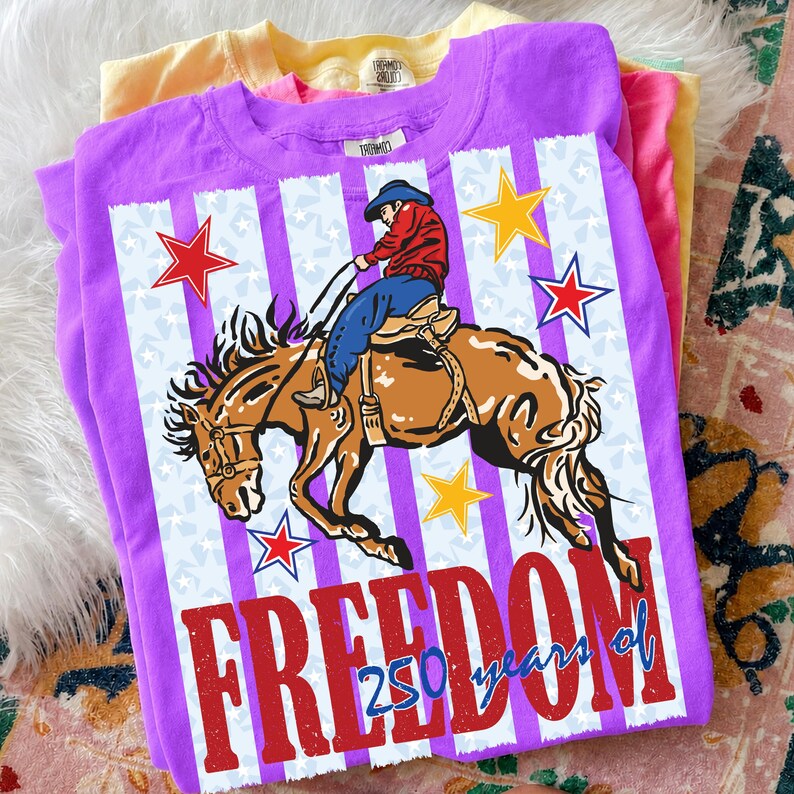 May include: A purple short-sleeve t-shirt featuring a graphic of a cowboy riding a bucking horse. The design includes red and yellow stars, vertical white stripes, and the red text "FREEDOM" with "250 years of" written in blue script below.