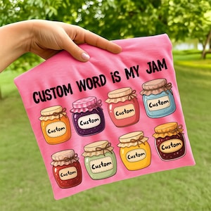 以下が含まれることがあります： 「CUSTOM WORD IS MY JAM」の黒い文字がプリントされたピンク色のTシャツ。各瓶に「Custom」の文字が入った、8つのジャム瓶のイラストが特徴です。瓶の色はオレンジ、紫、赤など様々です。