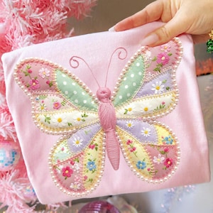 Pode incluir: Um item de tecido rosa com uma grande borboleta bordada. A borboleta tem asas de patchwork em cores pastel, adornadas com padrões florais e contornadas com pérolas falsas. As antenas são delicadas e o corpo é rosa texturizado.