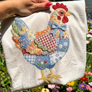 May include: A decorative kitchen towel featuring a patchwork chicken design. The chicken has a red comb and wattle, a blue bow, and wings with floral patterns. The towel is cream-colored with a garden background.
