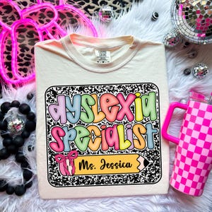 May include: Cream-colored t-shirt with a colorful graphic that reads "Dyslexia Specialist Ms. Jessica" in bubble letters. The graphic is framed by a black and white composition notebook design. A pink checkered tumbler is visible on the right.