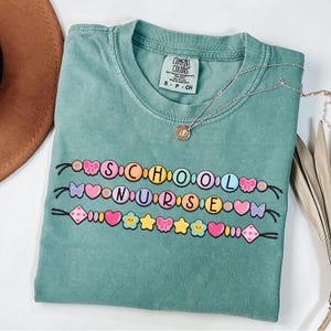 Könnte beinhalten: Ein türkisgrünes T-Shirt mit dem Schriftzug "SCHOOL NURSE" in einem farbenfrohen Perlendesign. Das Shirt hat einen Rundhalsausschnitt und kurze Ärmel. Eine silberne Halskette mit einem kleinen Anhänger liegt auf dem Shirt.