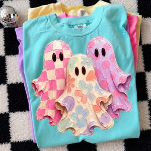 Pu&ograve; includere: Una t-shirt turchese brillante con una grafica di tre fantasmi con effetti di pittura materica a rilievo. Il fantasma a sinistra ha un motivo a scacchi rosa e crema, quello centrale fiori pastello e quello a destra pois rosa.