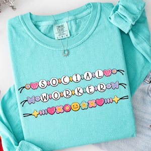 Pode incluir: Uma camisa turquesa de manga comprida com as palavras "SOCIAL WORKER" escritas em um design de contas. O design inclui corações, flores e carinhas sorridentes. Um colar de prata com um pingente de sol também é visível.