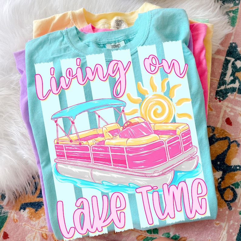 May include: A stack of pastel-colored t-shirts. The top shirt is light blue with a graphic of a pink and yellow pontoon boat, the text "Living on Lake Time", and a yellow sun. Other shirts are yellow, pink, and purple.