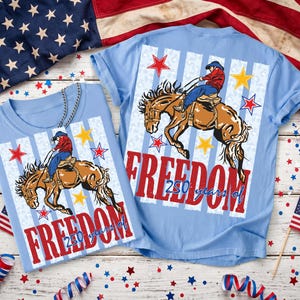 May include: A light blue t-shirt featuring a graphic of a cowboy riding a bucking horse. The design includes red and yellow stars, vertical stripes, and the red text "FREEDOM" with "250 years of" written in blue script below the main word.
