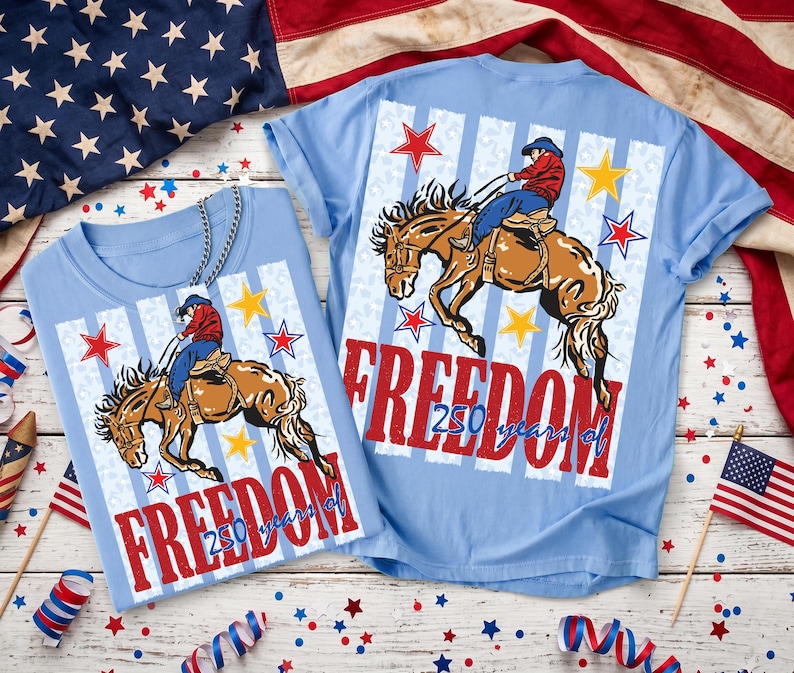 May include: A light blue t-shirt featuring a graphic of a cowboy riding a bucking horse. The design includes red and yellow stars, vertical stripes, and the red text "FREEDOM" with "250 years of" written in blue script below the main word.