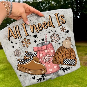 May include: A light gray tote bag featuring the text "All I need Is" in a whimsical font. The design includes a Dr. Pepper can with a pink bow, a brown knit hat, and brown shoes with a black and white checkered pattern.