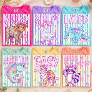 Peut inclure: Six t-shirts colorés avec des motifs fantaisistes. Chaque chemise présente un graphisme animalier différent et une phrase ludique : "MOOD," "SILLY," "LATER," "SHAKE THAT," "EASY," et "MESSY BESTIE." Les chemises sont roses, violettes, bleues, vertes, jaunes et oranges.