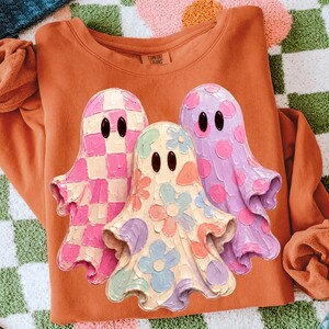 Pu&ograve; includere: Una felpa girocollo color arancio bruciato con una grafica raffigurante tre fantasmi con occhi ovali neri. Il fantasma a sinistra ha un motivo a scacchi rosa e crema, quello centrale una stampa floreale pastello e quello a destra pois viola.